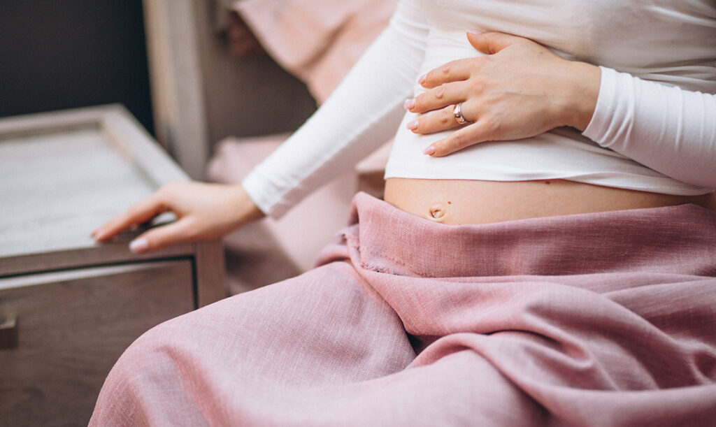 Schwangerschaft - Frau die am Bett sitzt und die Hand auf ihren Babybauch legt